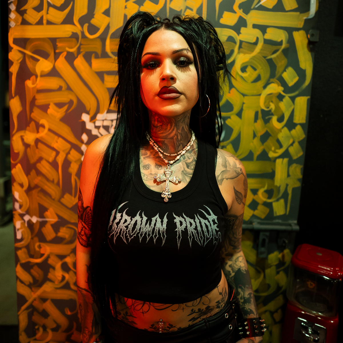 A tattooed woman with long black hair stands in front of a graffiti-covered yellow and black backdrop, wearing a black cropped tank top with the words 'CROWN PRIDE' in a stylized font.