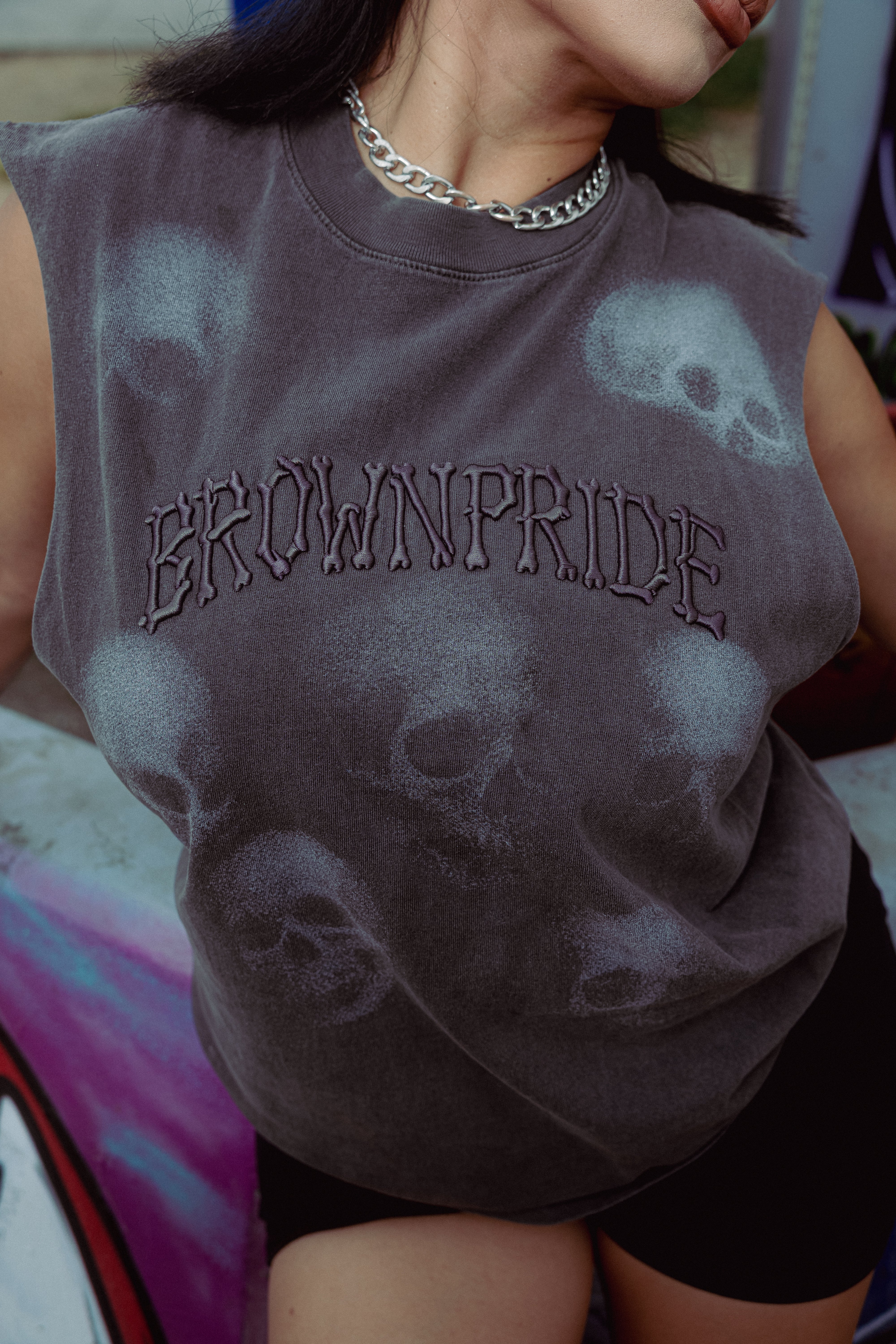 A close-up of a person wearing a sleeveless dark charcoal gray t-shirt with a mineral-wash finish, featuring a faded skull pattern and the word 'BROWNPRIDE' across the chest in a gothic font. The person is also wearing a silver chain necklace and black shorts.